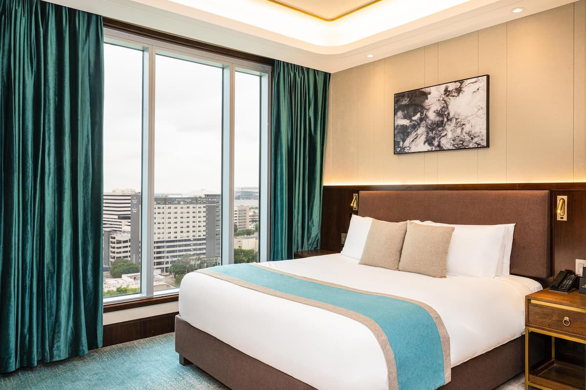 A hotel room with a large bed, a nightstand, a wall-mounted phone, and a city view through large windows.