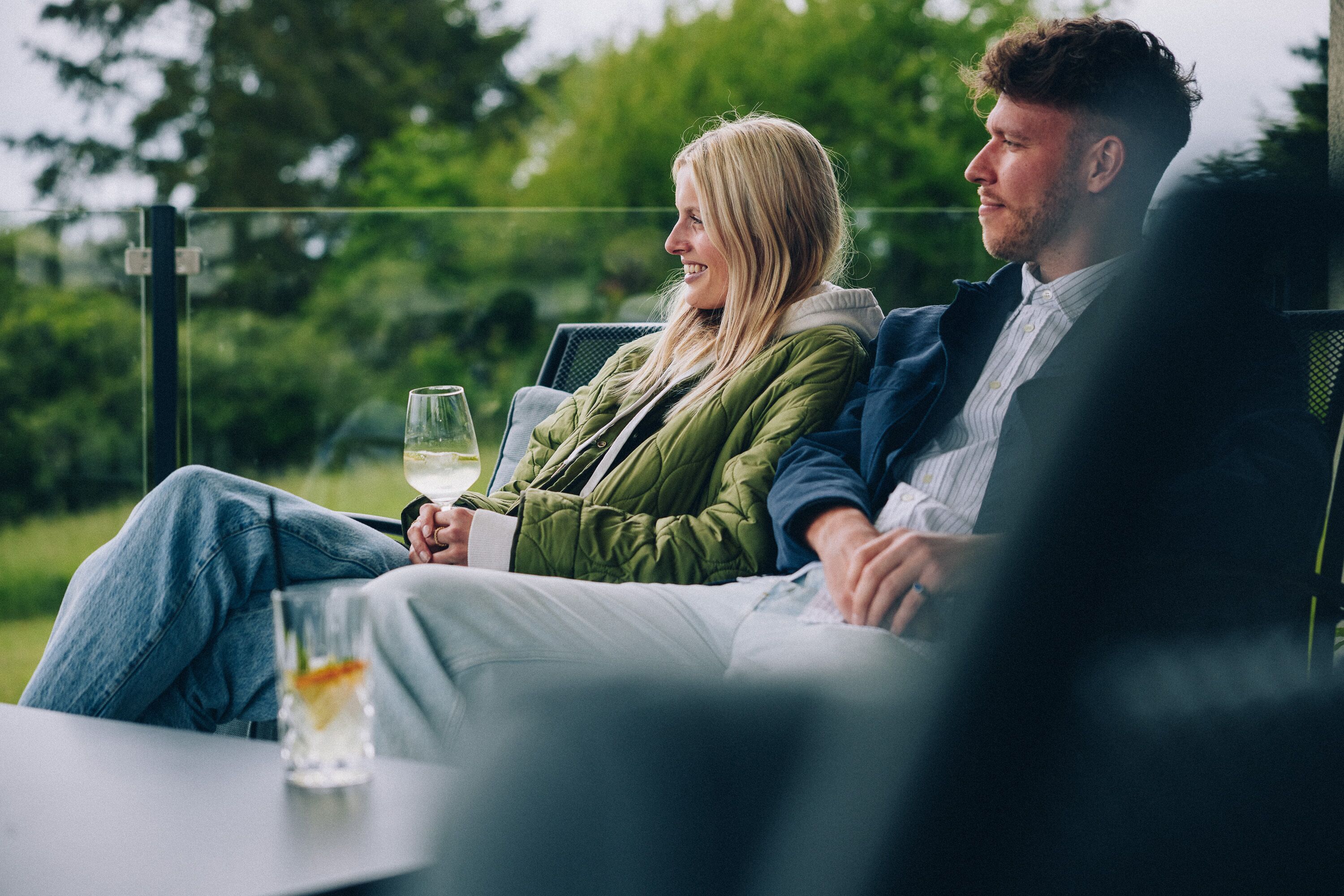 Two people sitting on a bench outdoors with a glass of wine in front of them.
