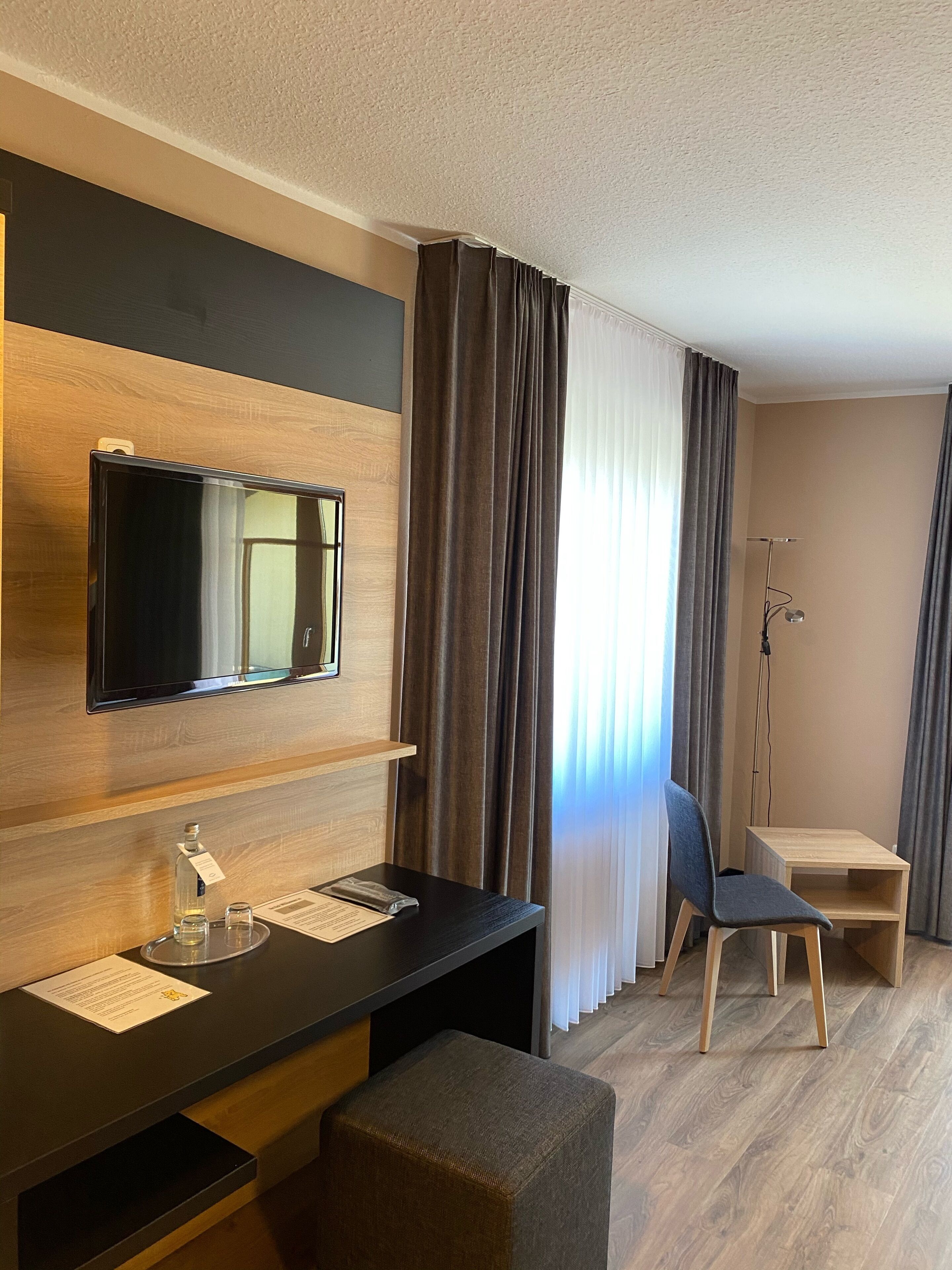 A modern hotel room with a wooden wall, a flat-screen TV, a desk with a chair, a small stool, and a standing lamp.