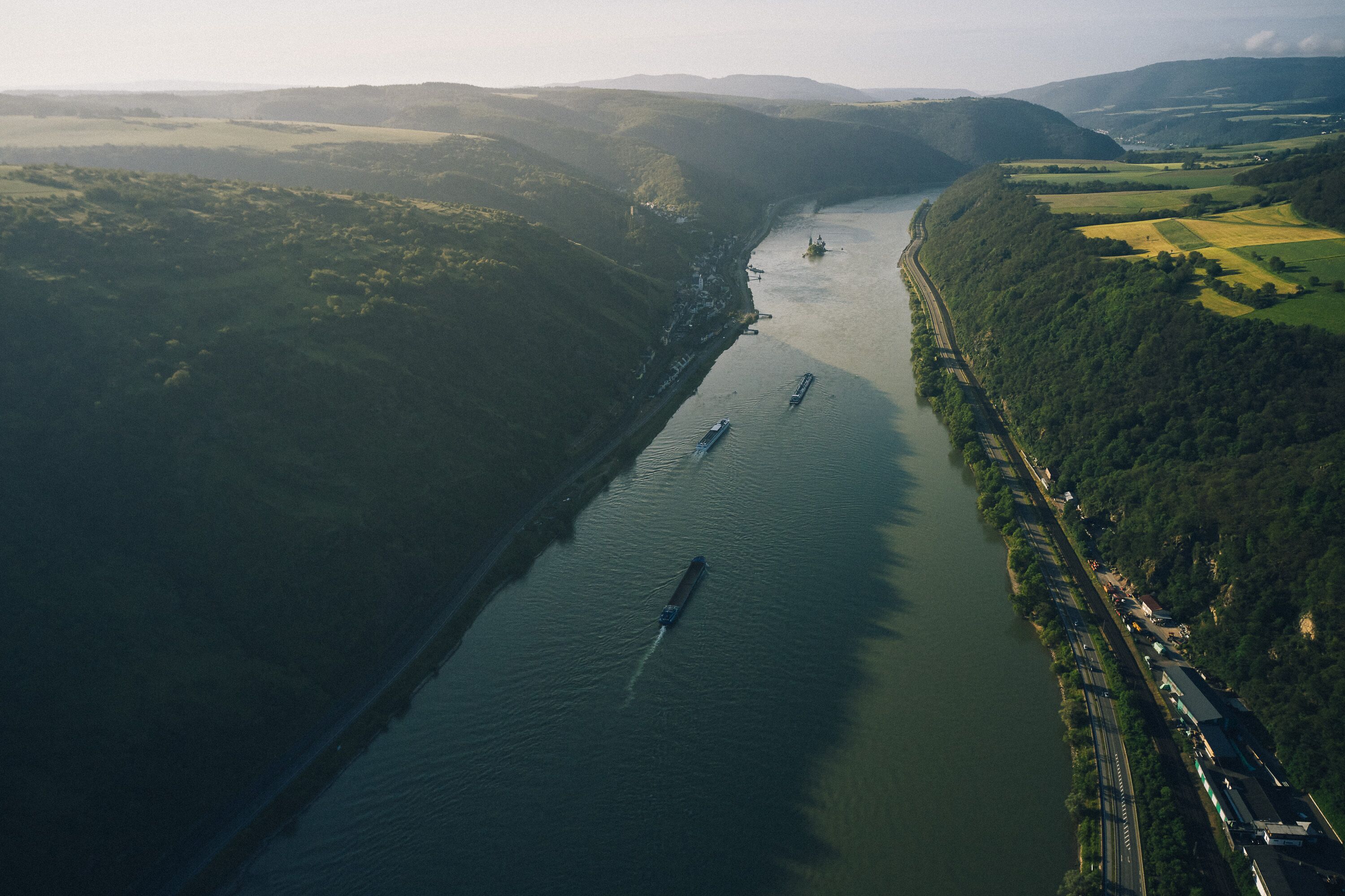 An aerial view of a river with boats, surrounded by hilly terrain and a bridge.