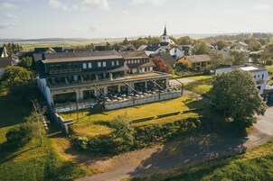 Un bâtiment à deux étages doté d’une terrasse et d’un espace extérieur aménagé pour s’asseoir, entouré de verdure et avec une église en arrière-plan.