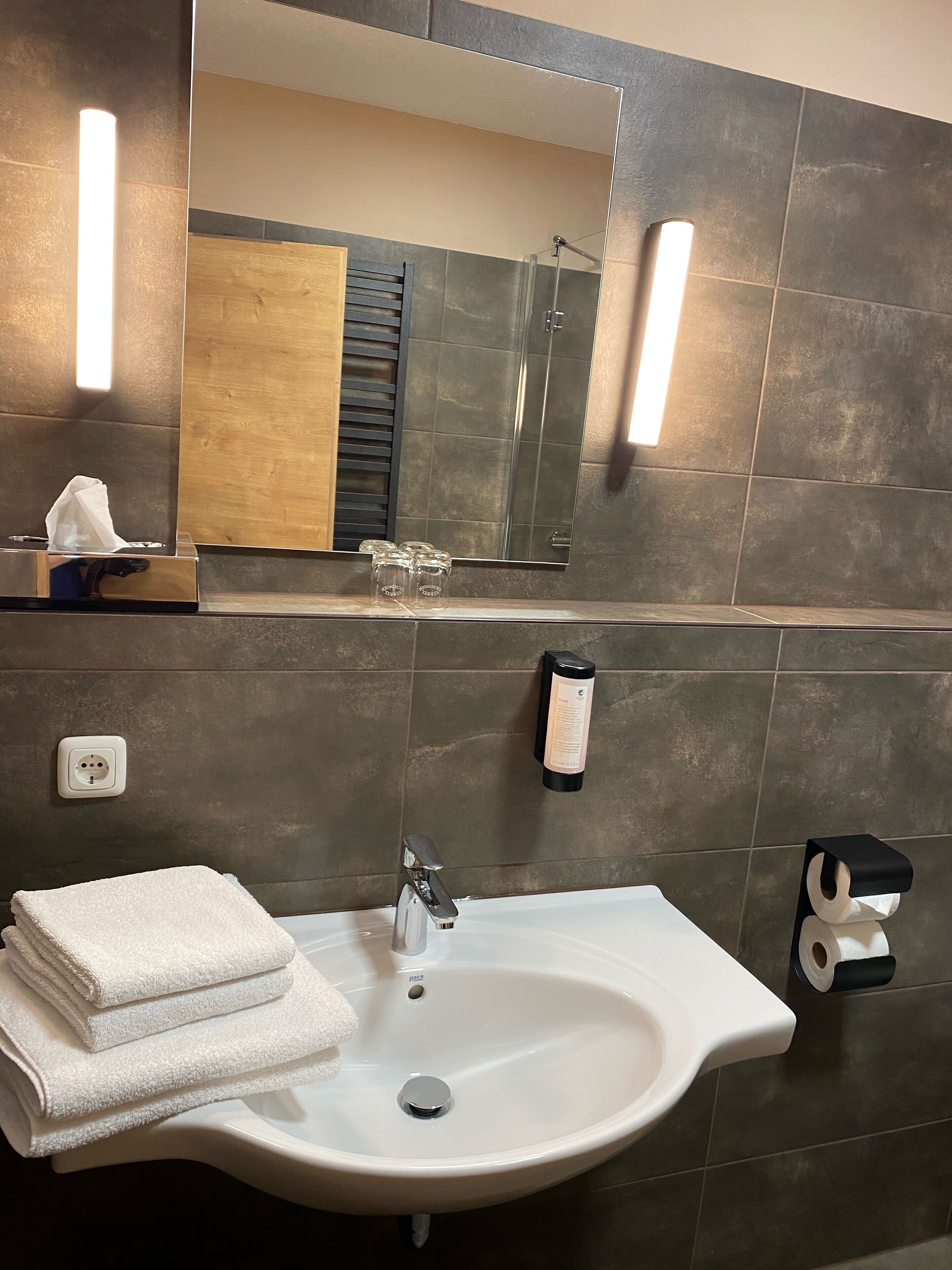 A bathroom with a sink, mirror, towel dispenser, and a shower area.