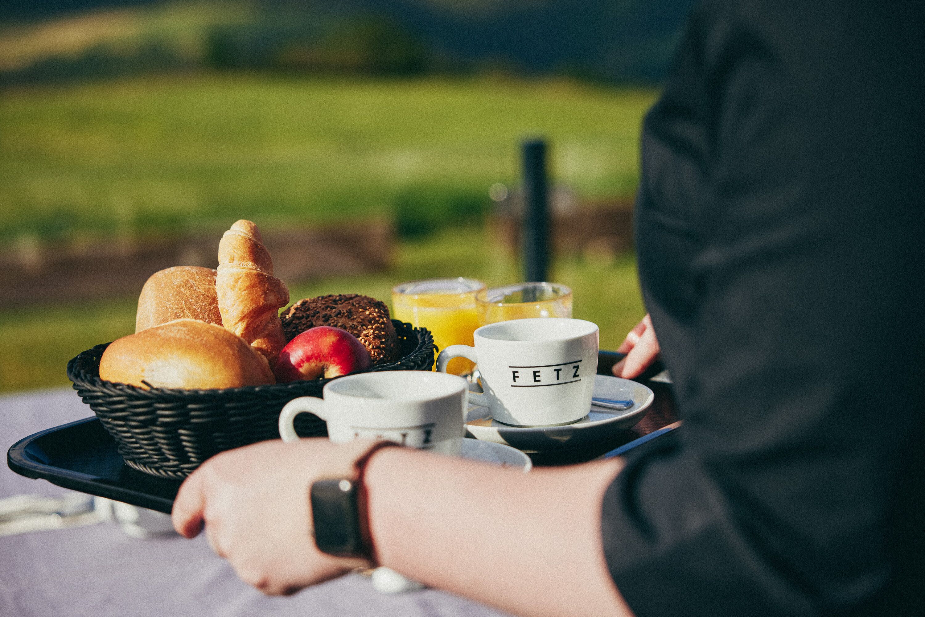 A breakfast tray with bread, fruits, and a mug labeled "FETZ" on a picnic table outdoors.