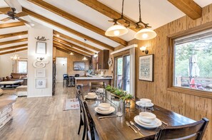 A spacious dining area with a wooden table set for four, a kitchen in the background, and a large window with a view of the outdoors.
