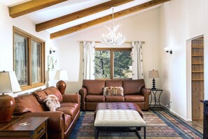 A spacious living room with a brown leather sofa, a white ottoman, and a chandelier. There are two windows with curtains, a wooden ceiling, and a rug on the floor.