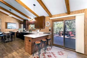 A modern kitchen with wooden beams, a large island with stools, a dining area with a view of the outdoors, and a sliding glass door leading to a deck.