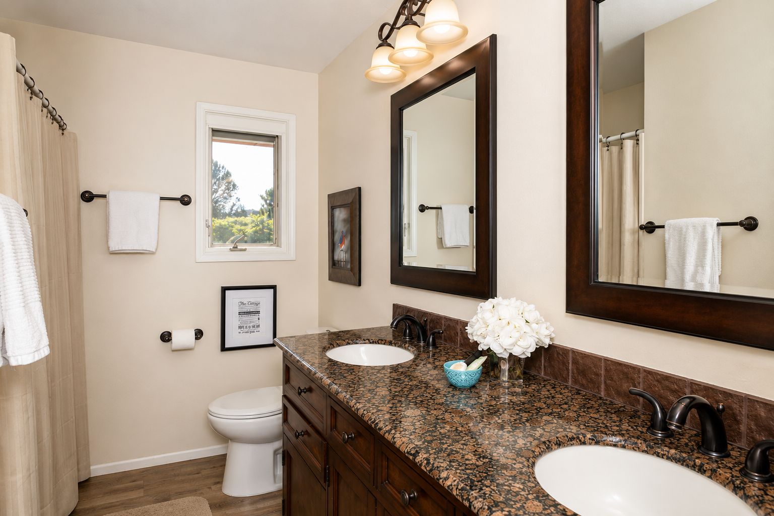 A bathroom with a double sink, a toilet, a framed picture, and a window.