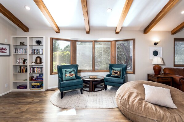 A living room with a wooden ceiling, a bookshelf, two armchairs, a round coffee table, and a large window.