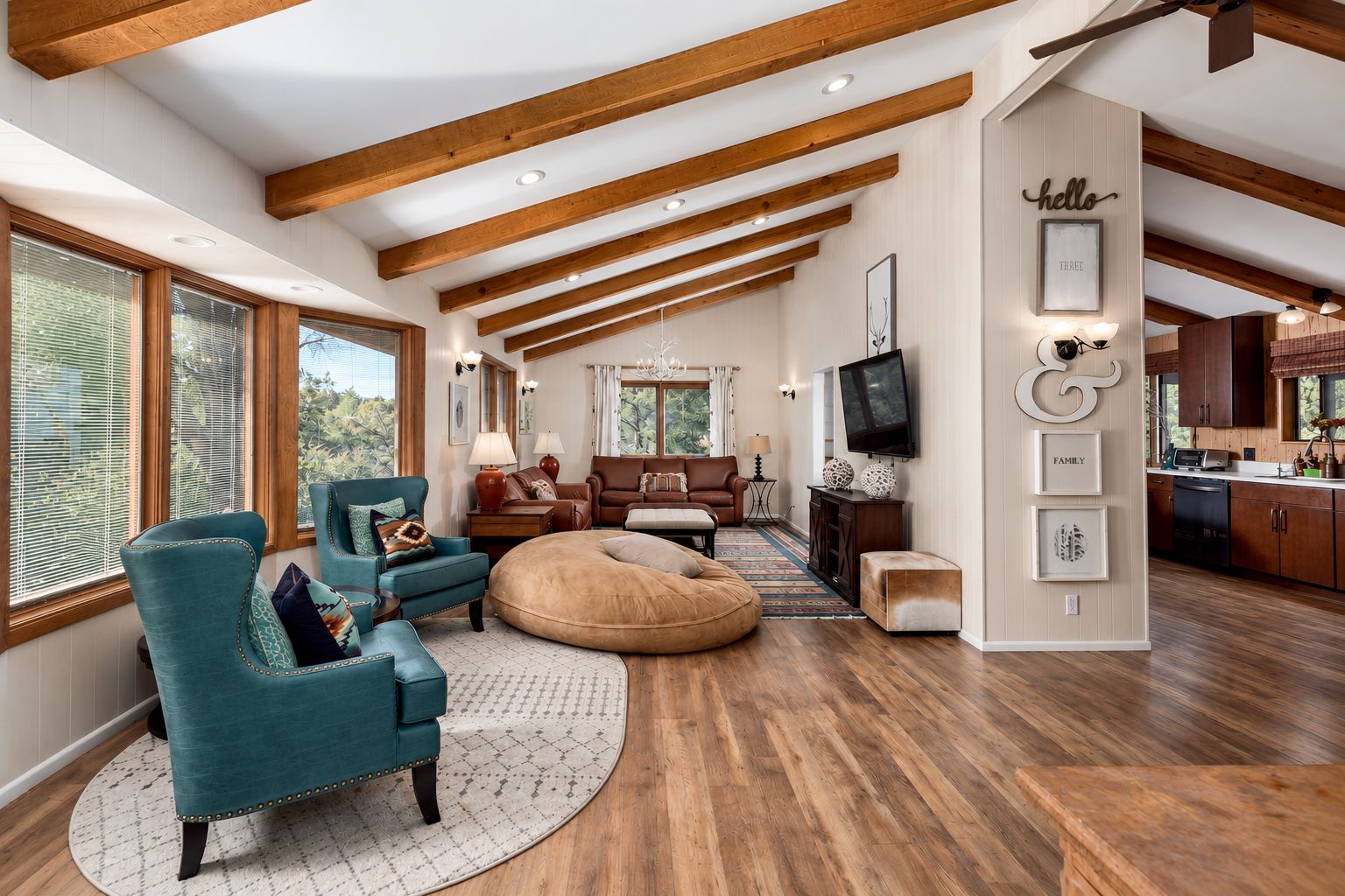 A spacious living area with a high ceiling, wooden beams, a large window, and a kitchen with wooden cabinets.