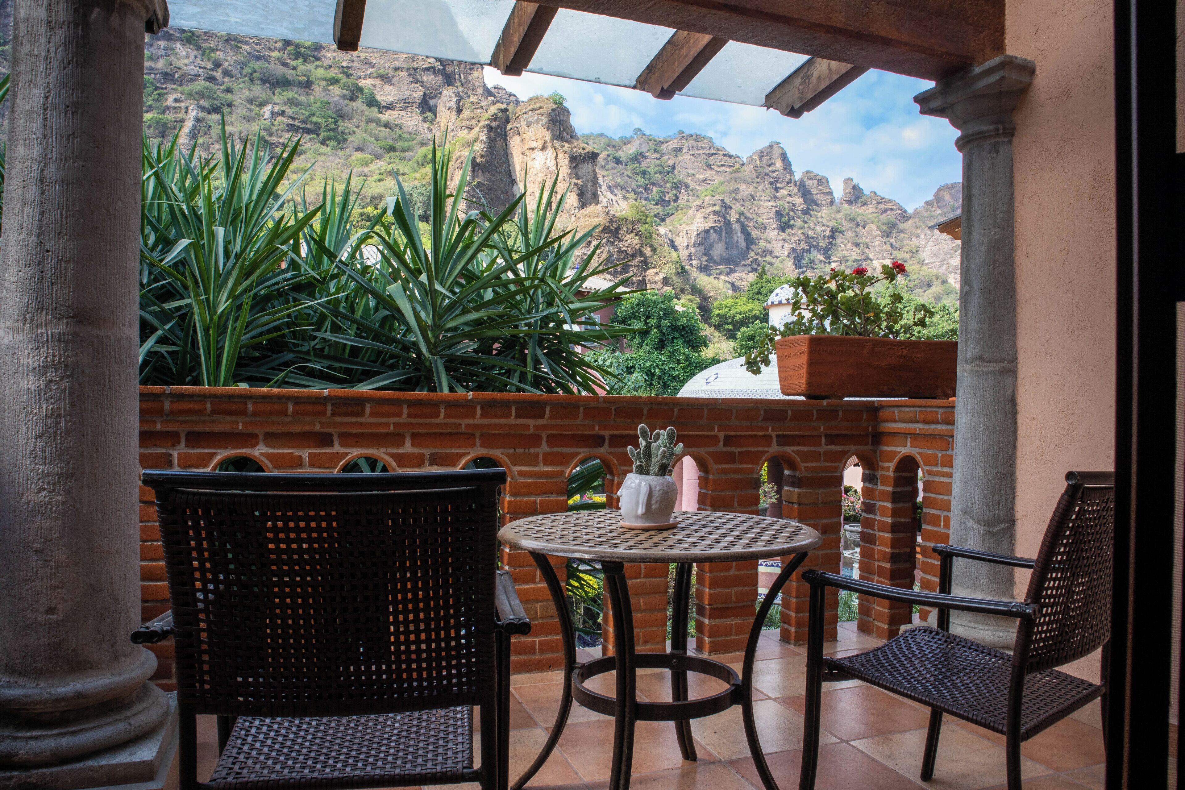 Eine Terrasse mit zwei Korbstühlen und einem runden Tisch, mit Blick auf eine gebirgige Landschaft, eine Backsteinmauer und Kübelpflanzen.