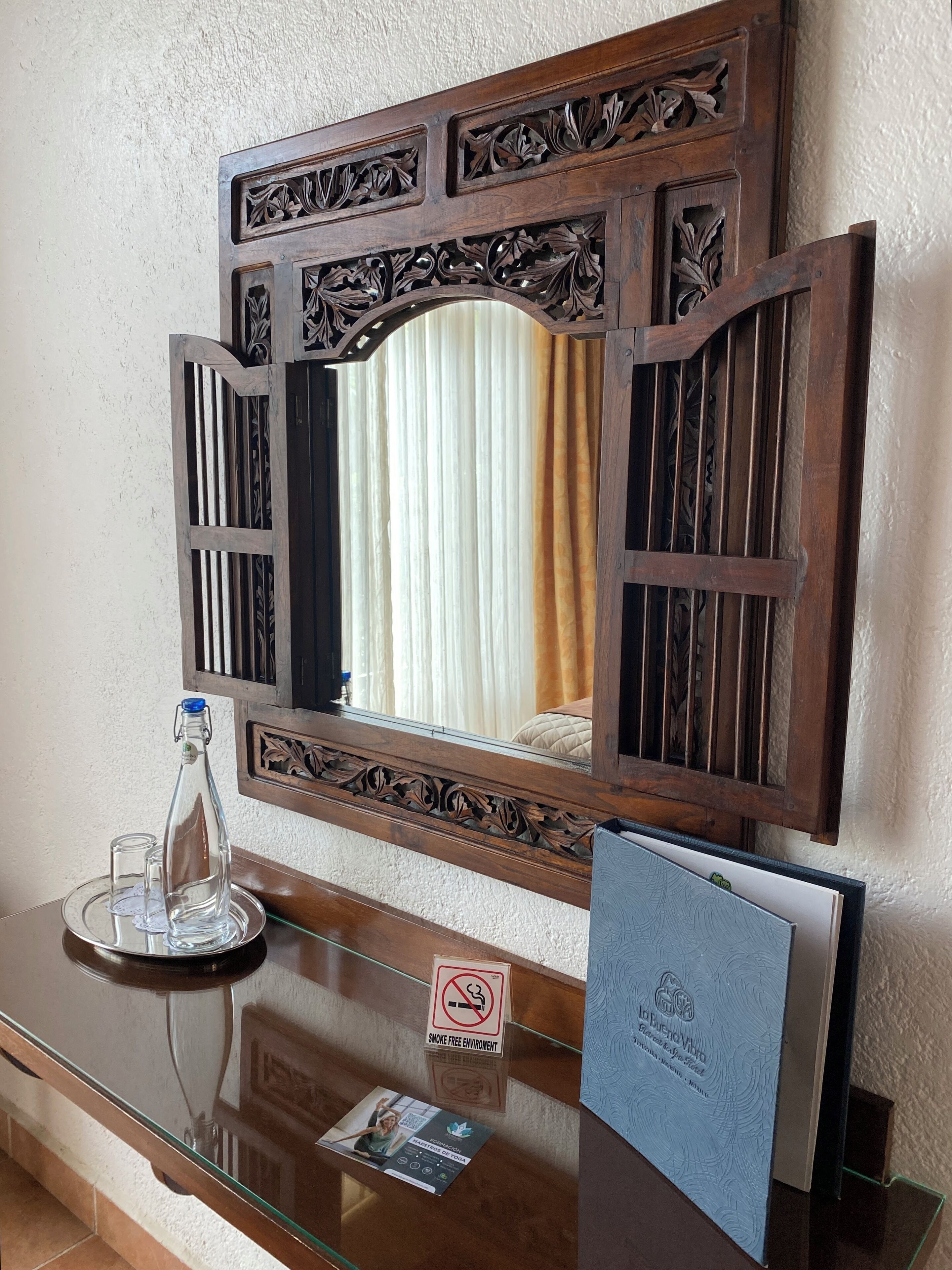 A wooden-framed mirror with intricate carvings, a glass table with a bottle and a glass, and a 'Smoke Free Environment' sign.