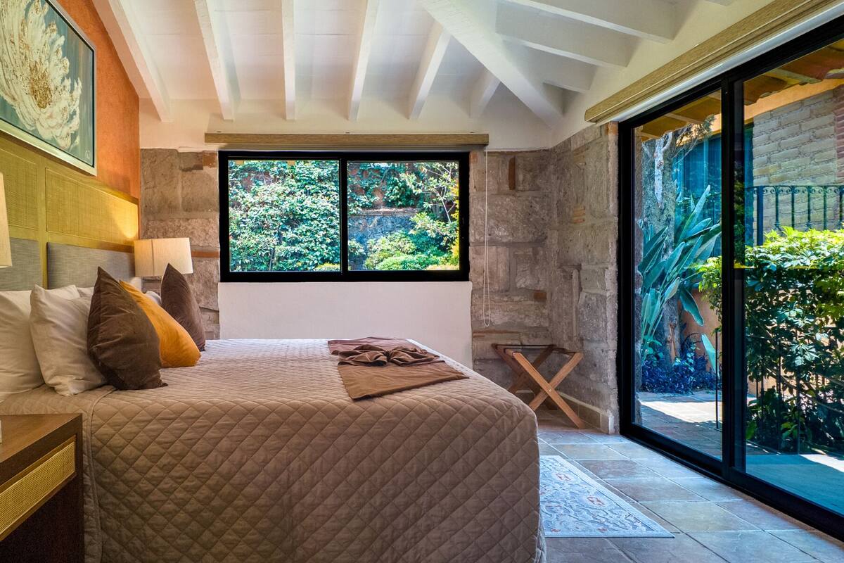 A bedroom with a large bed, a nightstand, a lamp, and a window with a view of greenery.