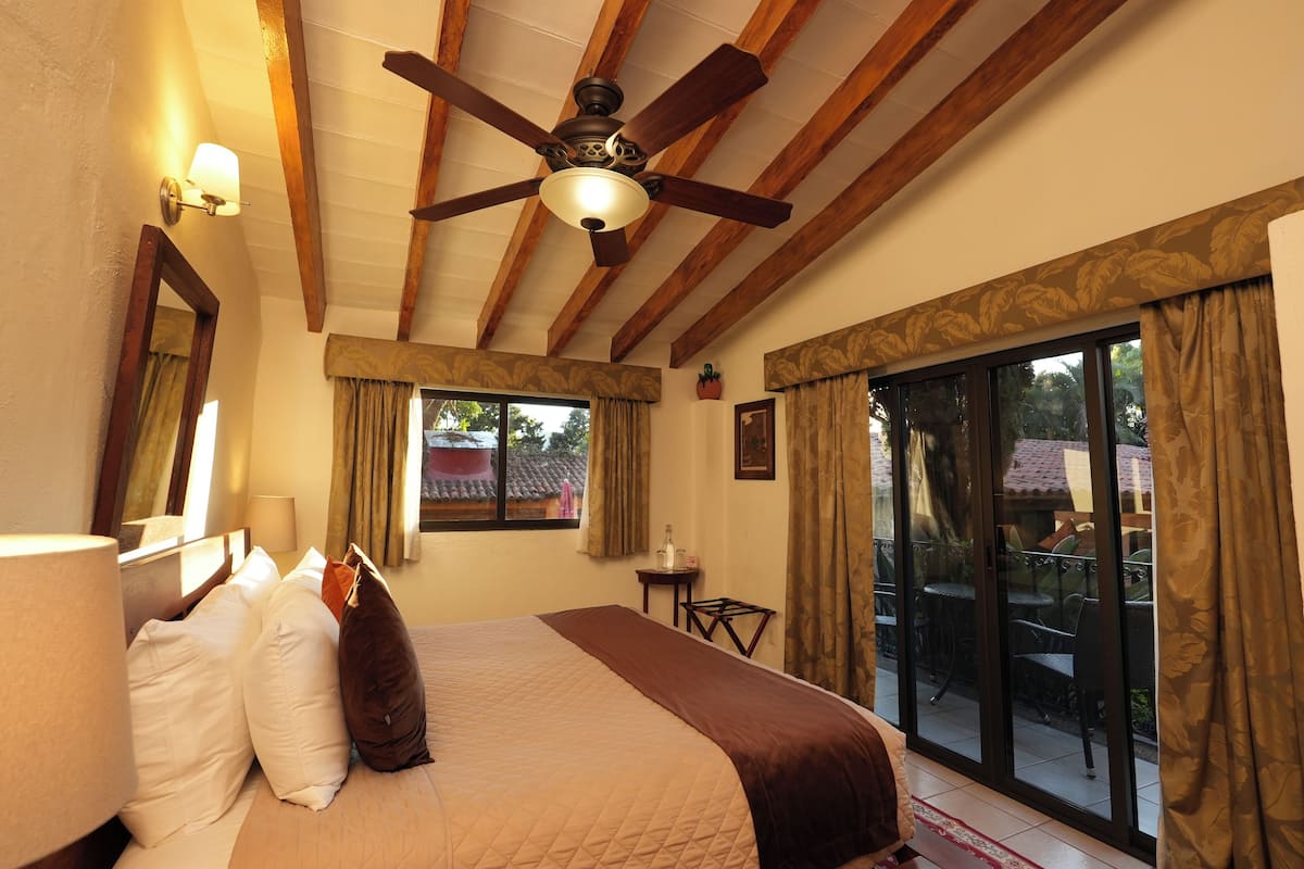 A hotel room with a bed, ceiling fan, large window with curtains, and a view of the outside patio.