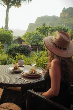 Una persona sentada en una mesa al aire libre con platos de comida y una copa de vino, con vistas a un jardín exuberante y un paisaje montañoso.