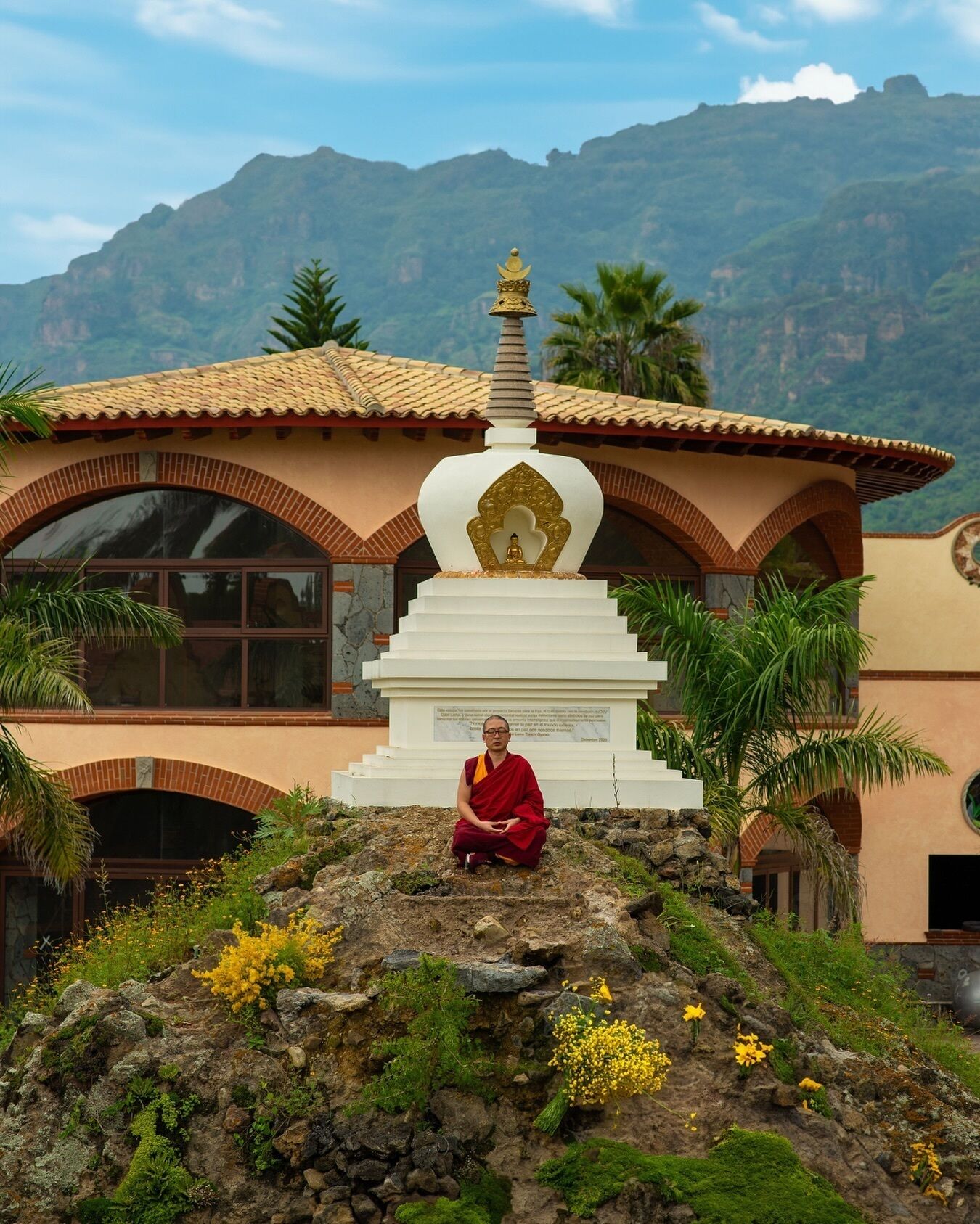 A person sitting on a rock in front of a building with a large white stupa and a mountain in the background.