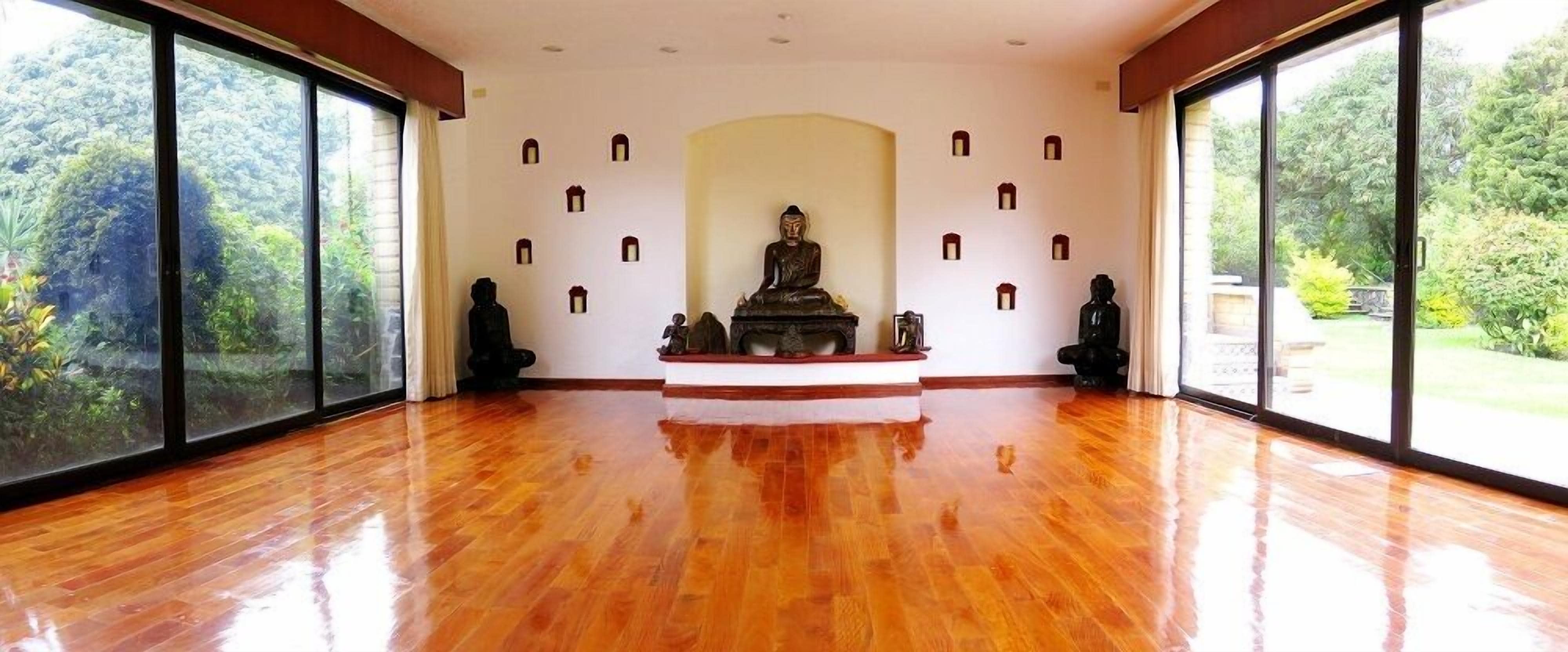 A spacious room with a polished wooden floor, large windows offering a view of greenery, and a central statue on a pedestal.