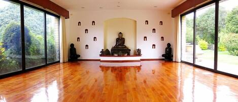 A spacious room with a polished wooden floor, large windows offering a view of greenery, and a central statue on a pedestal.