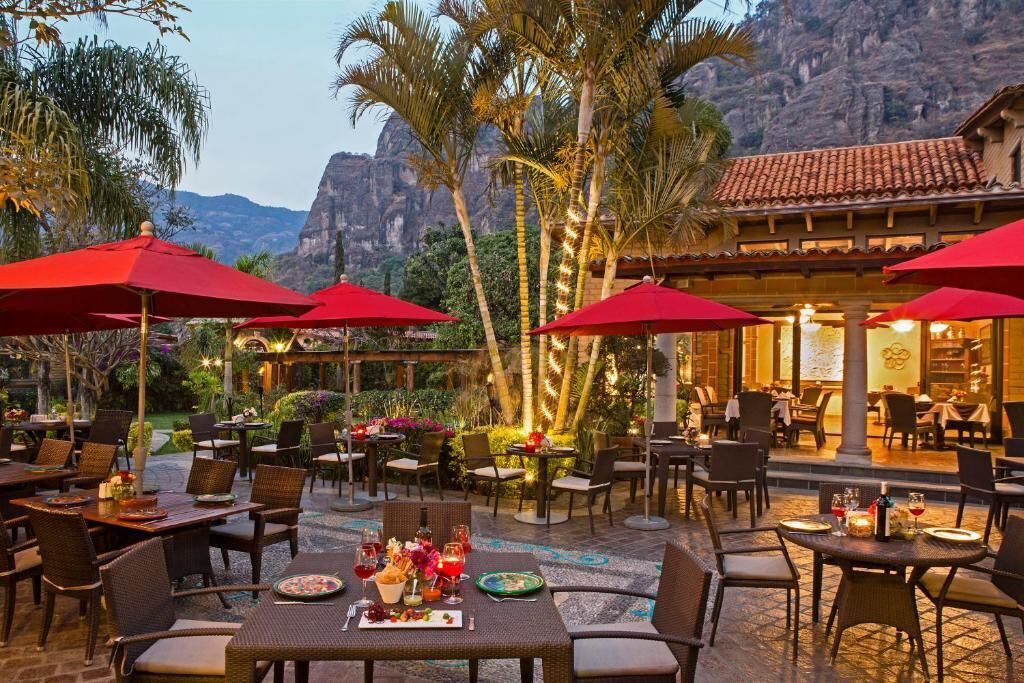 An outdoor dining area with tables and chairs, red umbrellas, and a view of mountains in the background.