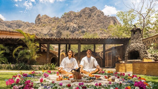 Two people meditating on a colorful flower arrangement in a courtyard with a mountain backdrop.