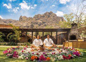 Two people meditating on a colorful flower arrangement in a courtyard with a mountain backdrop.