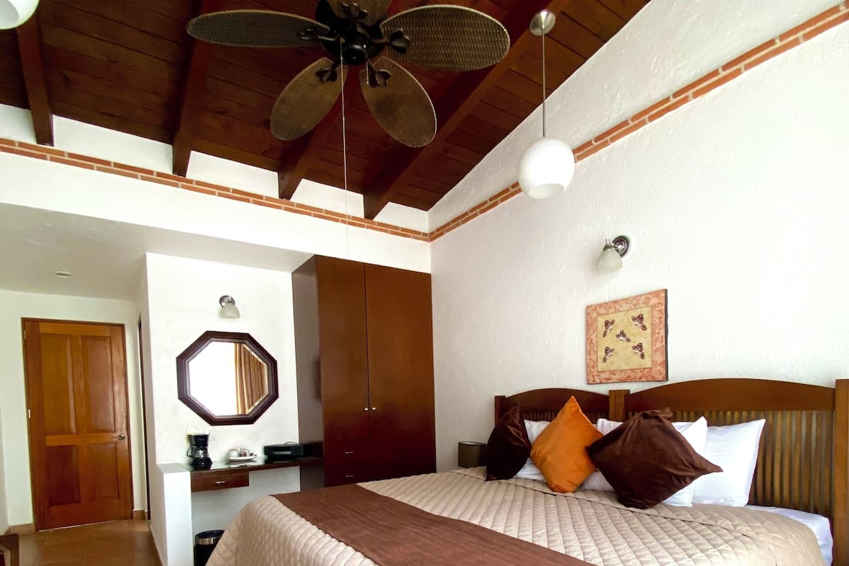 A bedroom with a large bed, a ceiling fan, a desk with a chair, a mirror, and a patterned rug.