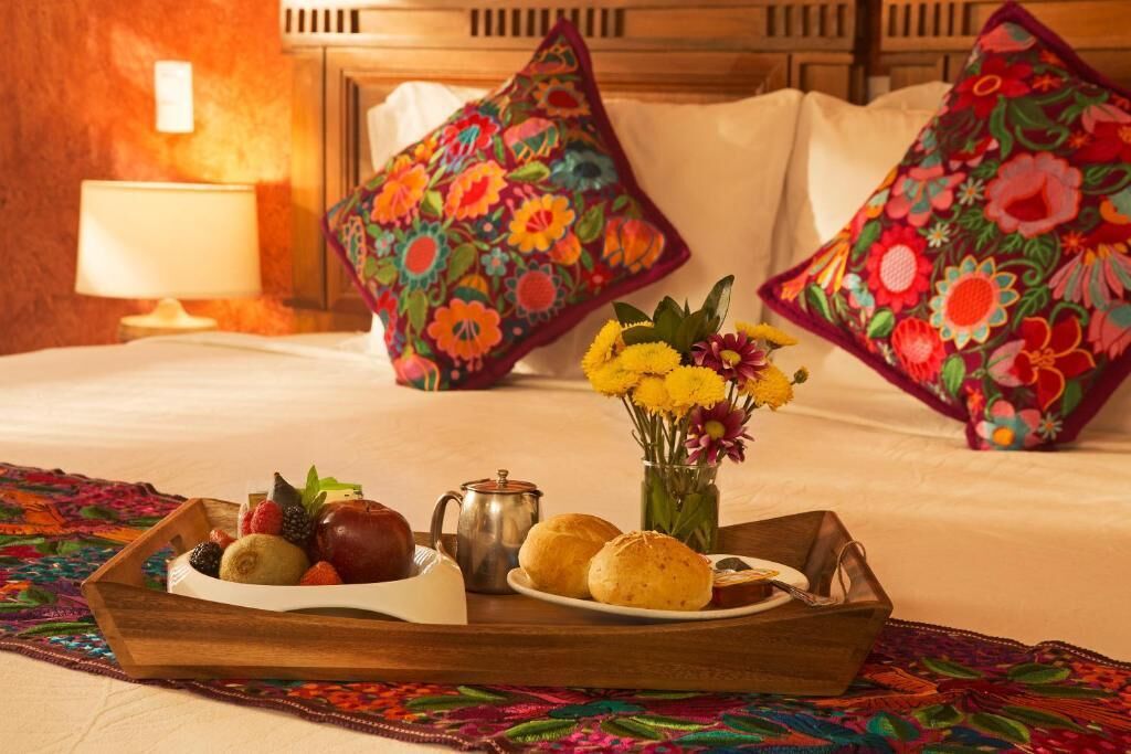 Una cama bien hecha con almohadas florales de colores, una bandeja con frutas, un juego de té y un florero con flores.