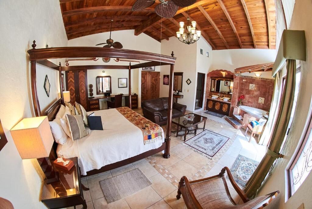 A spacious bedroom with a four-poster bed, a sitting area with a chair and table, and a view of a dining area with a chandelier.