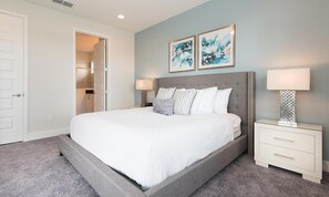Quarto com uma cama grande, duas mesas de cabeceira com abajures, uma porta e um corredor que dá para um banheiro.