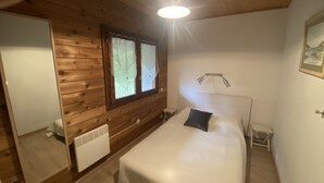 Een slaapkamer met een houten plafond, een bed met een witte matras en een donker kussen, een nachtkastje, een wandlamp en een raam met gordijnen.