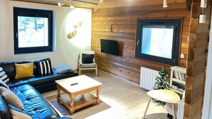 Een gezellige woonkamer met een houten plafond, een zwarte leren bank, een houten salontafel, een kleine witte tafel en een aan de muur bevestigde televisie.