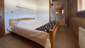 A bedroom with a wooden bed, a bedside table, a small stool, a white wall, a wooden ceiling, and a window with curtains. - GITE 2 to 5 PERS HAUT JURA PLEINE NATURE (Morbier)