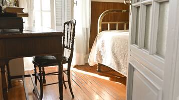 A bedroom with a wooden floor, a bed with a white bedspread, a chair, and a desk with books and a vase of flowers.