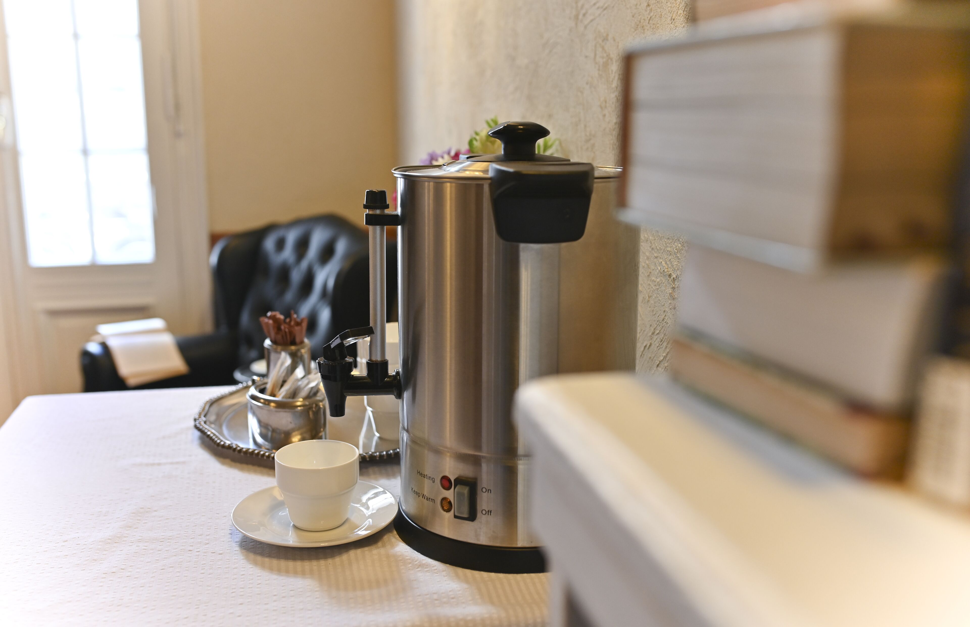 A coffee maker on a table with a cup and saucer, a tray, and a decorative item.