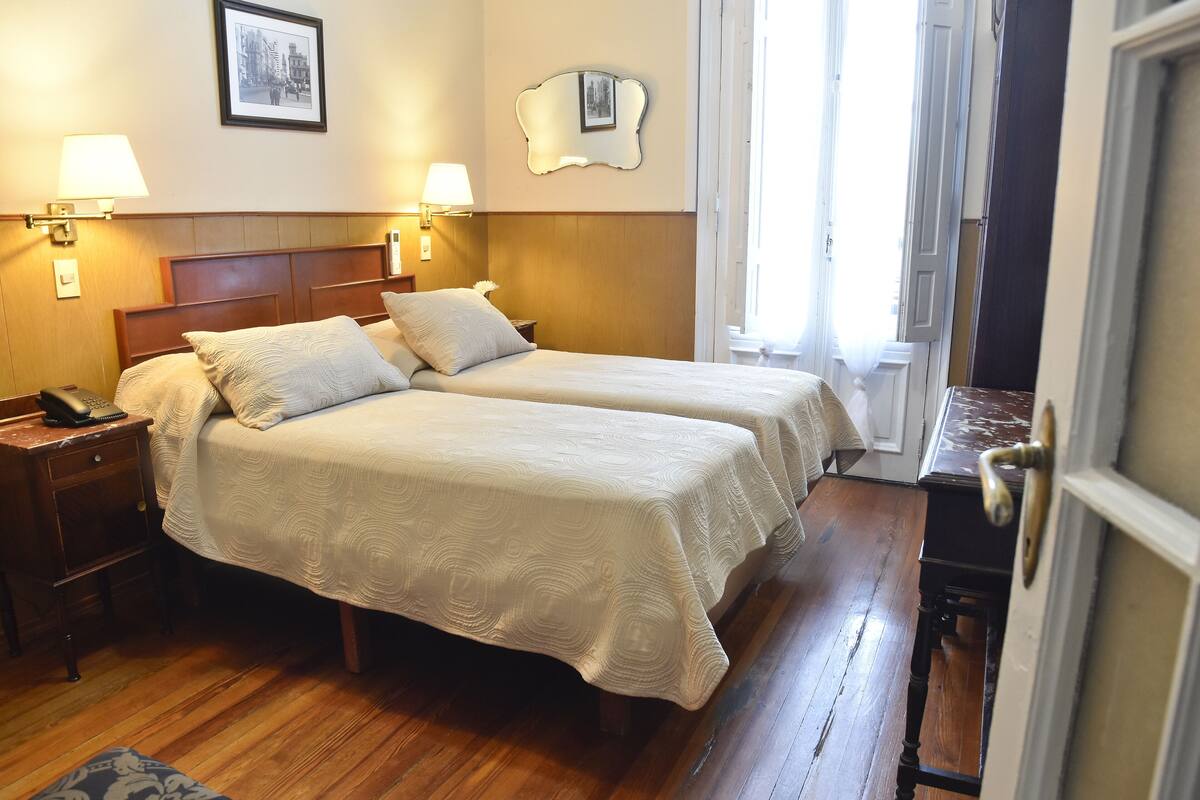 A hotel room with two beds, wooden floors, bedside lamps, a nightstand, and a framed picture on the wall.