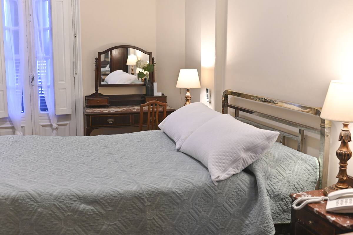 A neatly made bed with a patterned bedspread, a wooden nightstand with a lamp, a dresser with a mirror, and a chair.