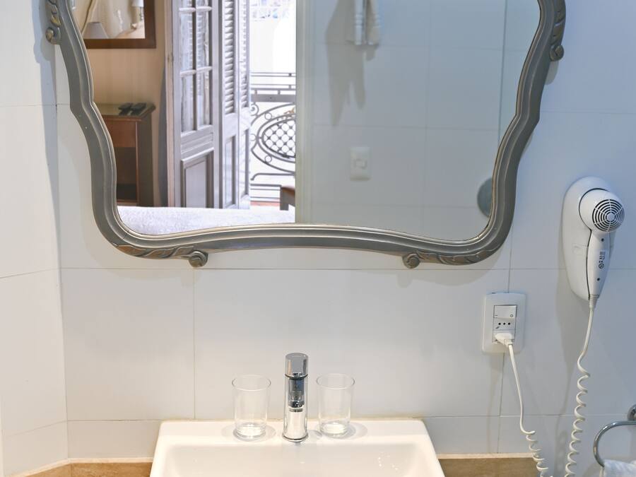 Un baño con un espejo grande y decorativo, un lavamanos blanco, dos vasos, un secador de pelo y un perchero para toallas.