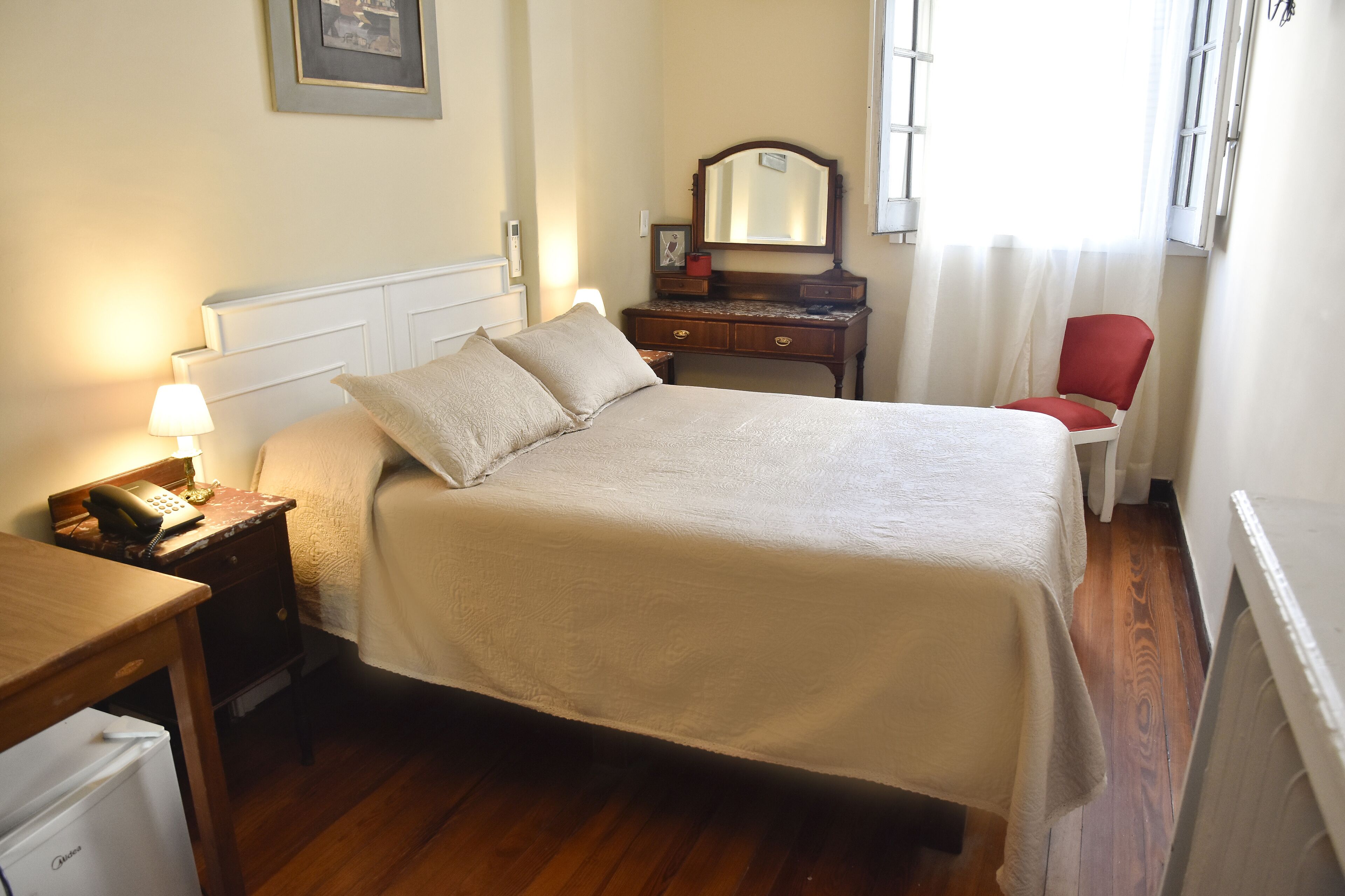 A neatly arranged bedroom with a bed, bedside table, lamp, dresser, mirror, and a red chair.