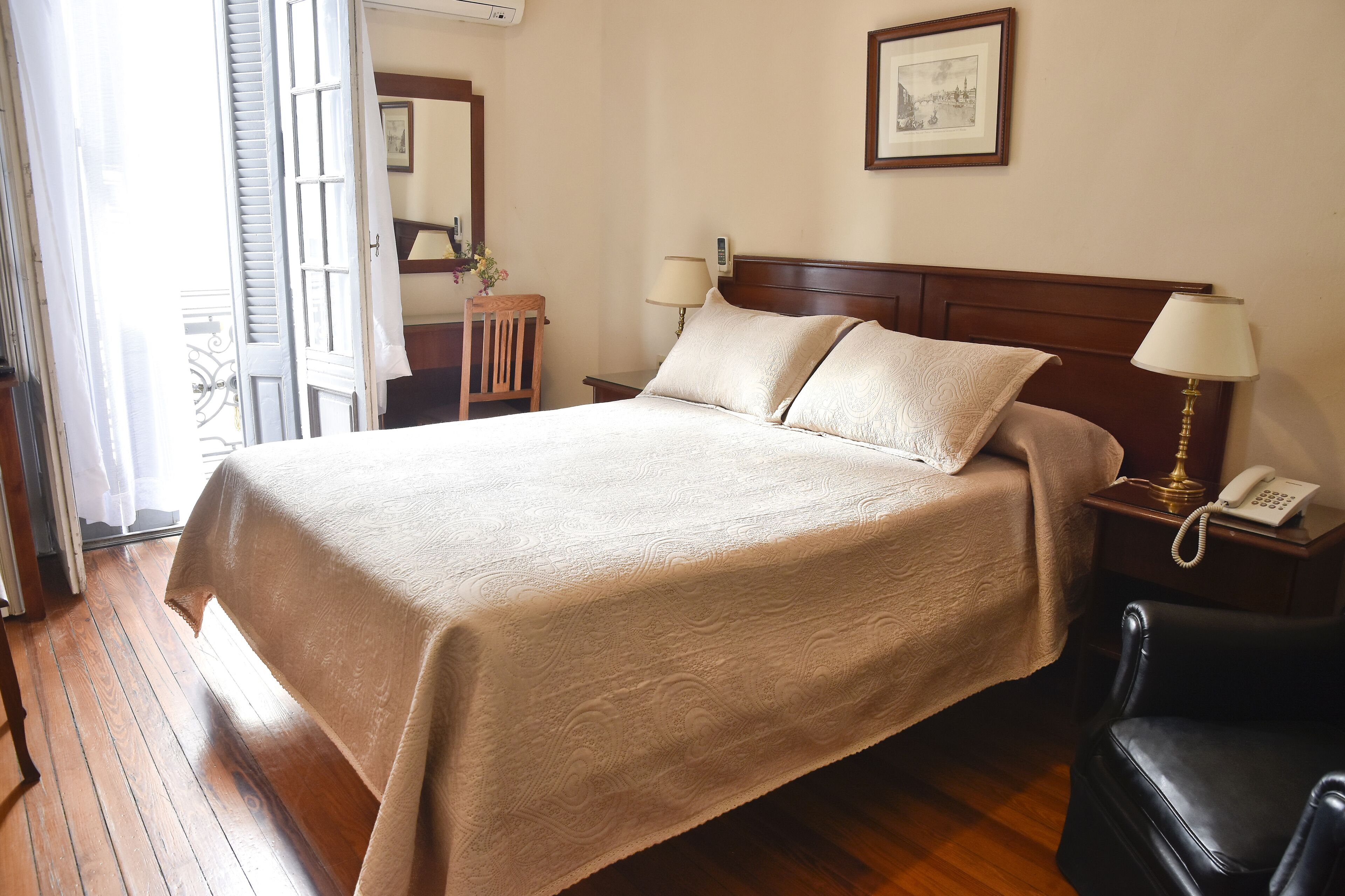 A hotel room with a large bed, two bedside tables with lamps, a wooden nightstand, a chair, and a door leading to a balcony.