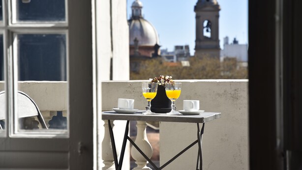 Un balcón con una mesa lista para dos personas, con un jarrón de flores y dos vasos de jugo de naranja.