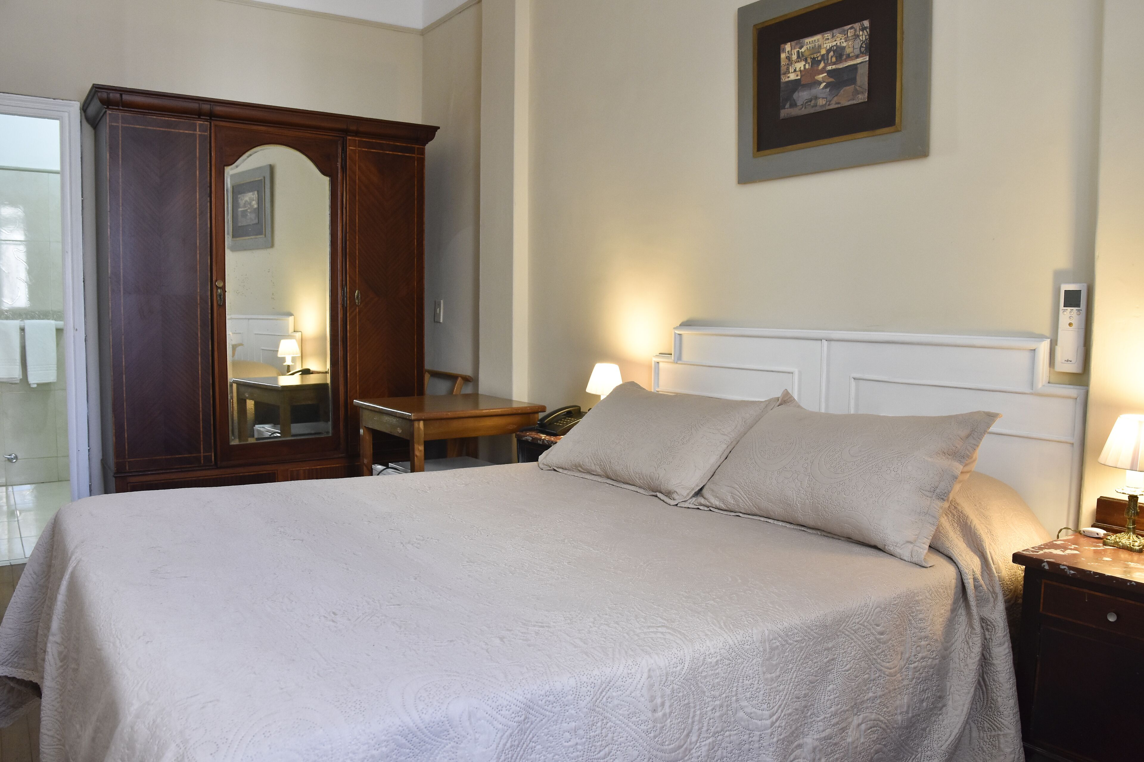 Un dormitorio ordenado con una cama grande, un armario de madera, un escritorio con una silla y una mesita de noche con una lámpara.