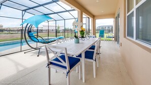 Un patio exterior techado con piscina, mesa blanca con cojines azules y una silla mecedora azul.
