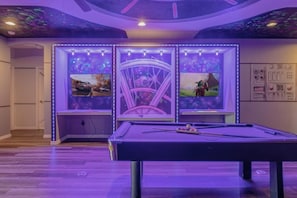 A pool table with LED lighting, a ceiling with a space-themed mural, and two framed screens displaying video games.