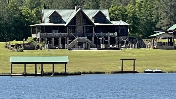 A large, multi-story lodge with a prominent porch and a covered deck area, situated by a lake with a well-maintained lawn and a small dock. - Lodge on The Lake (Rutledge)
