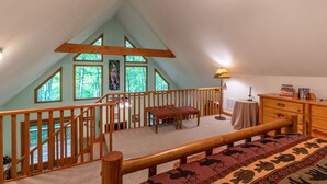 Ein gemütliches Schlafzimmer im Dachgeschoss mit schrägen Wänden, Holzbalken, einer kleinen Sitzecke und Blick auf den Wald durch große Fenster.