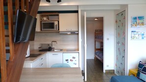 Une cuisine compacte équipée d’un four à micro-ondes, d’un évier et d’un petit réfrigérateur. Un téléviseur est fixé au mur, et on peut apercevoir un lit superposé dans la pièce adjacente.