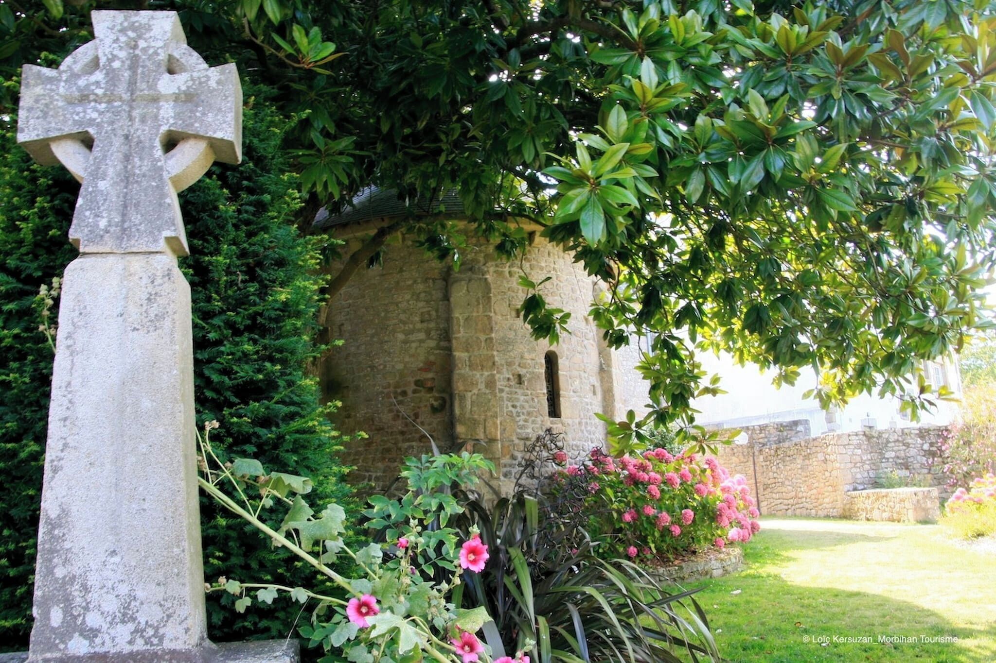 A stone cross in a garden with a stone wall and flowering plants.