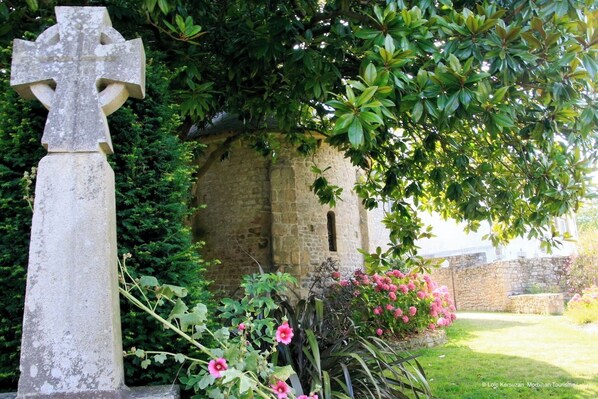 Une croix en pierre dans un jardin, avec un mur en pierre et des plantes en fleurs.