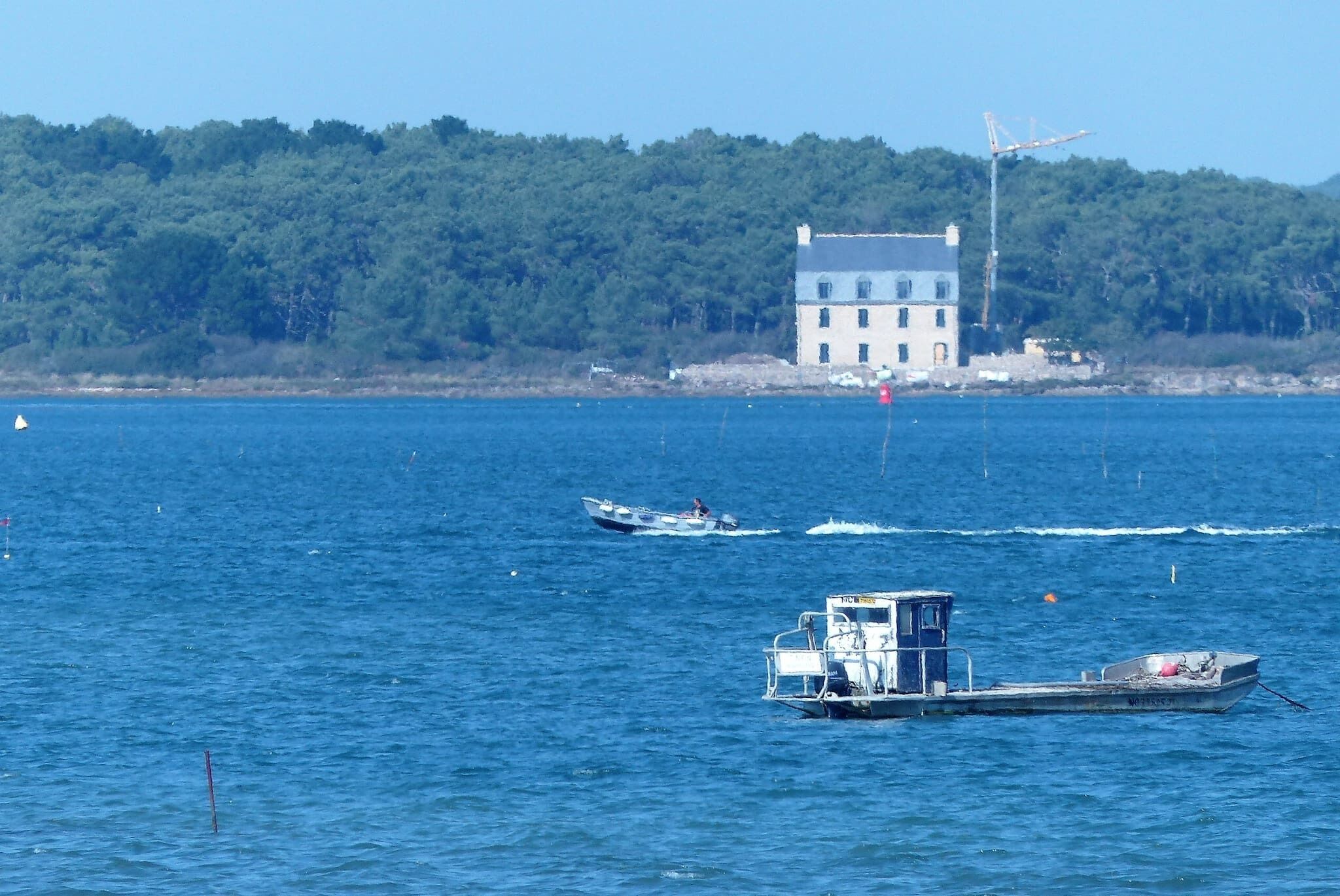 Un paysage côtier avec un grand bâtiment situé sur une colline, un ferry et un plus petit bateau sur l’eau.