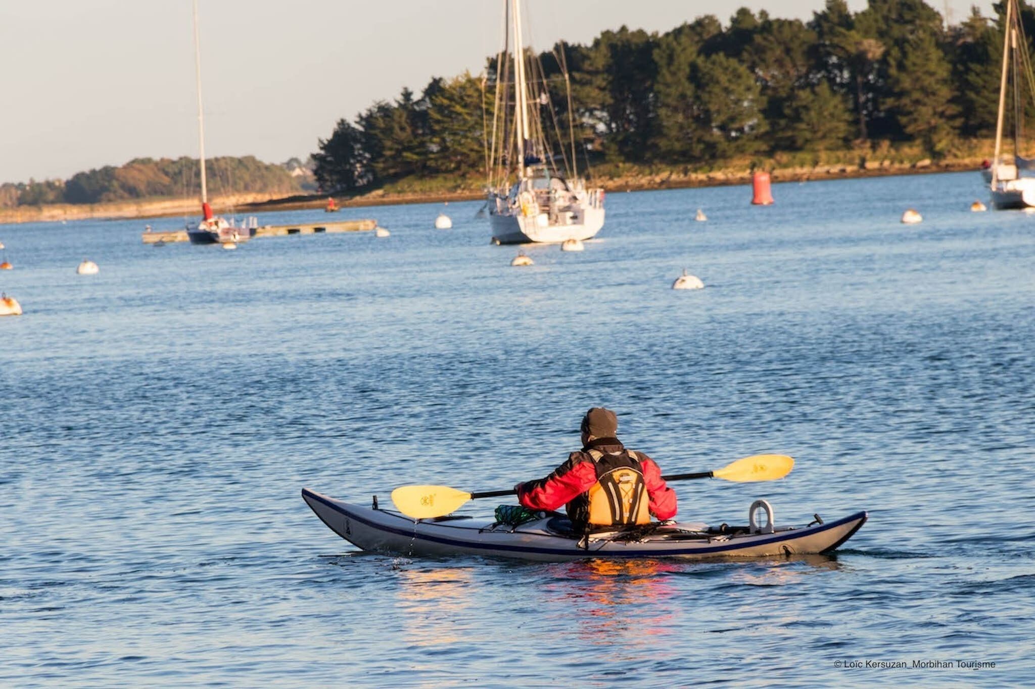 A kayaker in a small boat on a calm body of water with several sailboats in the background.