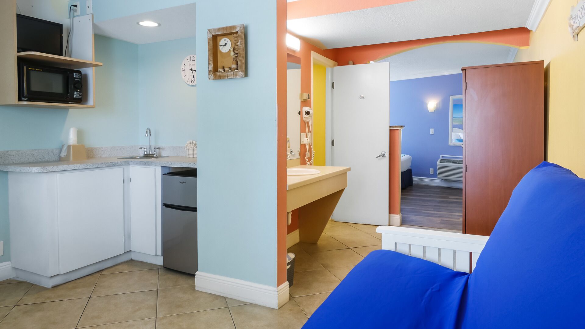 Een kleine keuken met een magnetron, spoelbak en een kleine koelkast. Er hangt een klok aan de muur en er is een deur naar een badkamer.